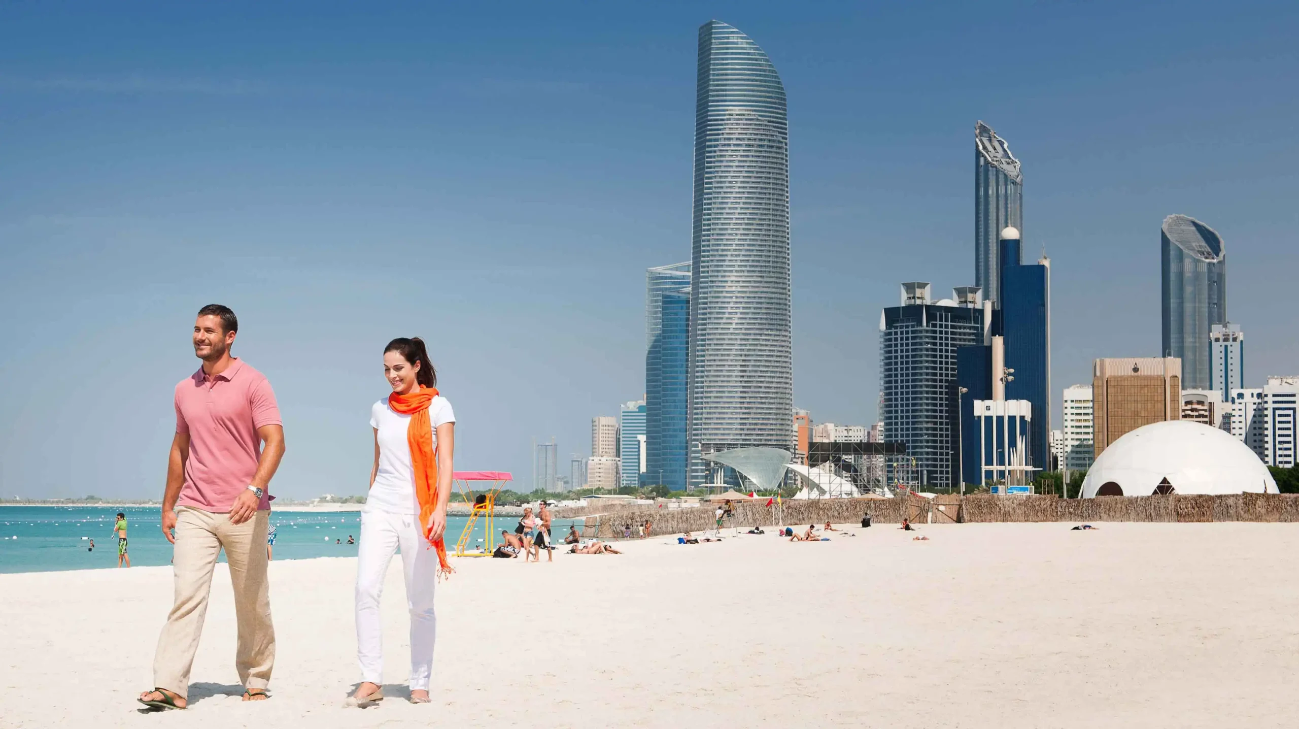 Couple walking along the white sand beach of Abu Dhabi Corniche with the city's skyscraper skyline in the background