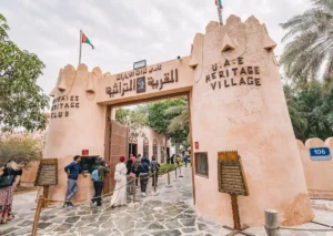 Entrance gate to Abu Dhabi Heritage Village with traditional mud-brick archway, Arabic and English signage, and visitors walking through