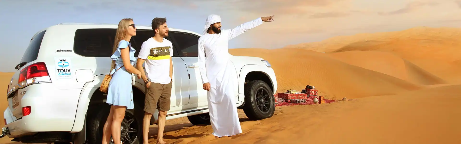 Guide pointing toward the desert horizon while two tourists stand beside a white 4x4 near a dune picnic setup