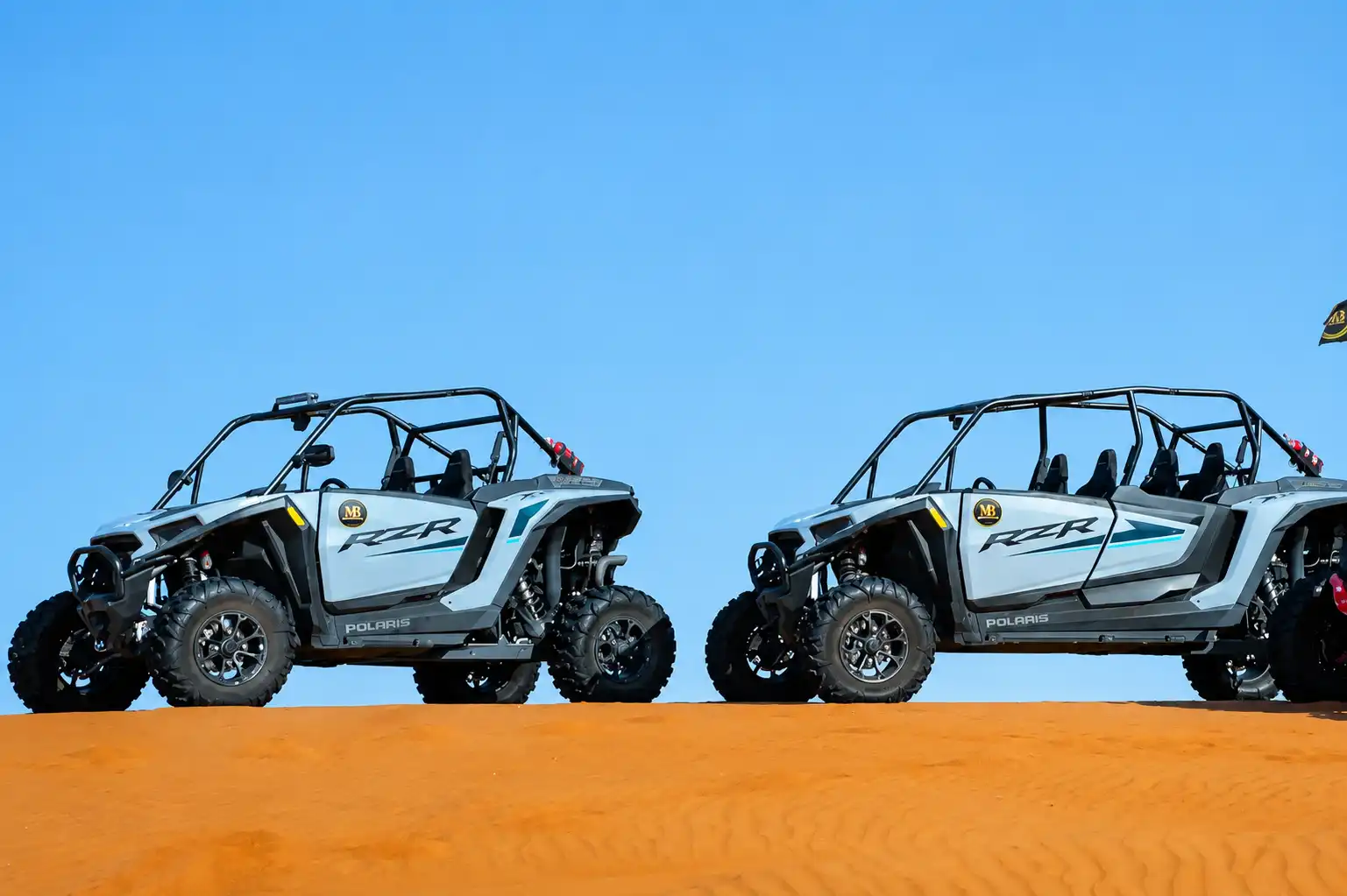 Two white Polaris RZR off-road buggies parked side by side atop a Dubai sand dune against a clear blue sky