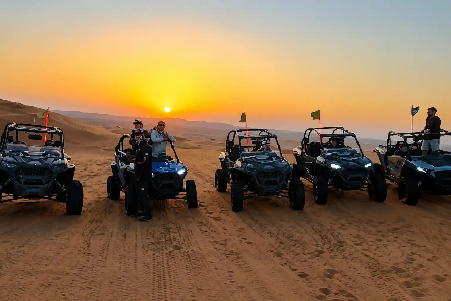 Row of off-road buggies lined up on a desert dune at sunset with guides standing beside them