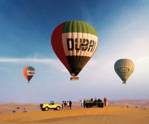Hot air balloons with 'Dubai' branding rising over the desert at sunrise, with a 4x4 vehicle and guests gathered at the launch site