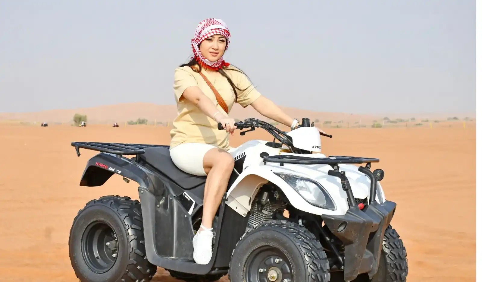 Woman wearing a traditional red-and-white keffiyeh riding a white quad bike ATV across the Dubai desert sand