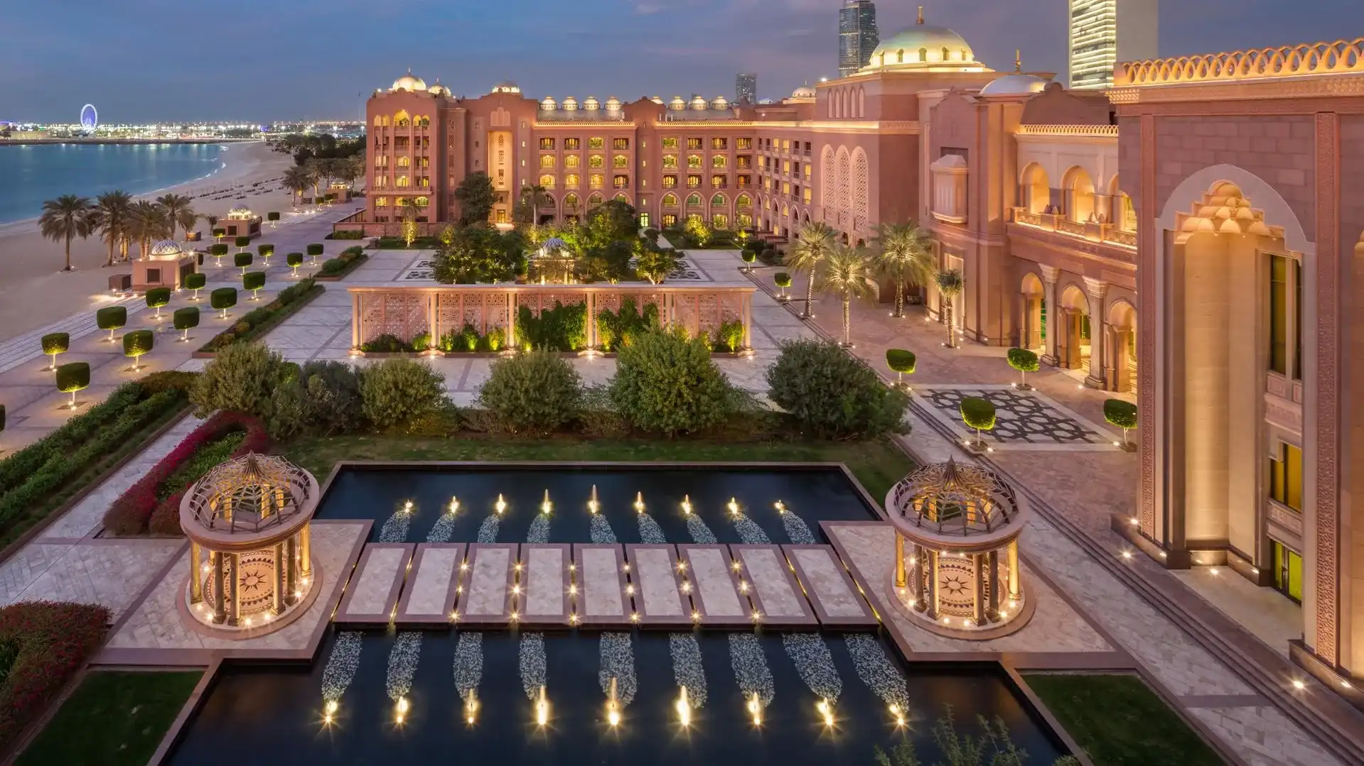 Emirates Palace hotel in Abu Dhabi illuminated at dusk, showing its golden domes, reflecting pools, and fountain-lined walkway