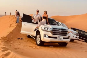 Couple posing on a white Toyota Land Cruiser during a morning desert safari in Dubai, with golden dunes and other 4x4 vehicles in the background