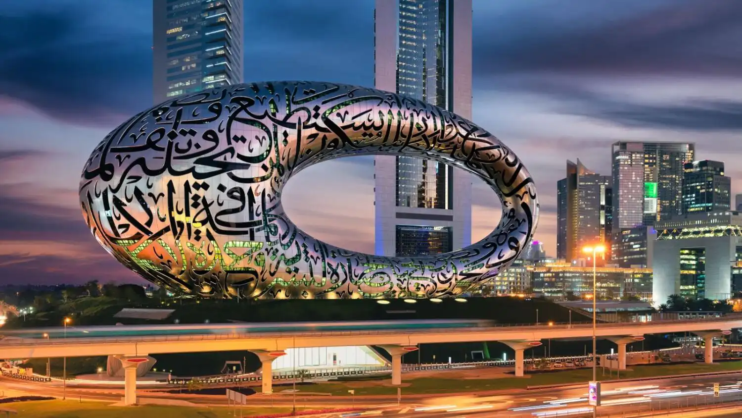 Museum of the Future's illuminated elliptical torus building in Dubai at dusk, covered in Arabic calligraphy, with city skyline and highway traffic trails below