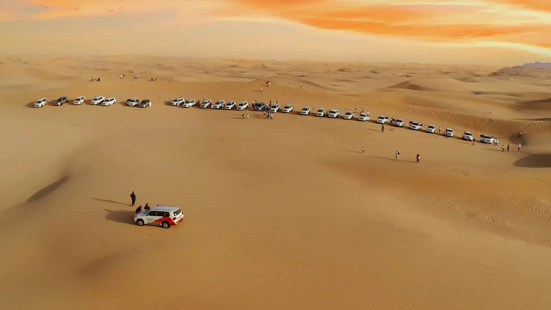 Aerial view of a long line of white 4x4 vehicles parked in the desert with tourists gathered at a sunset viewpoint