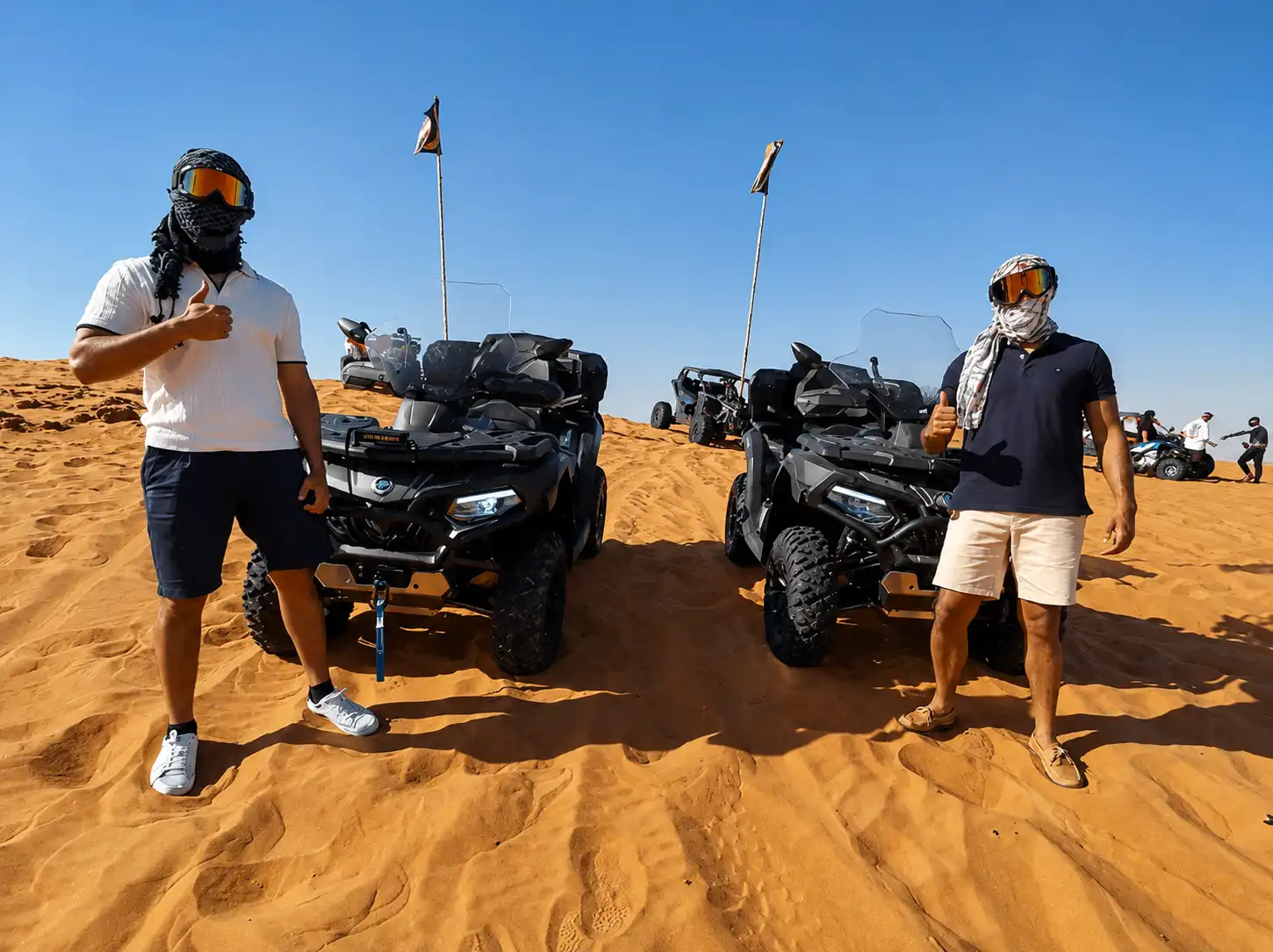 Two guests giving thumbs up beside their quad bikes in the Dubai desert, wearing traditional shemagh headscarves and goggles